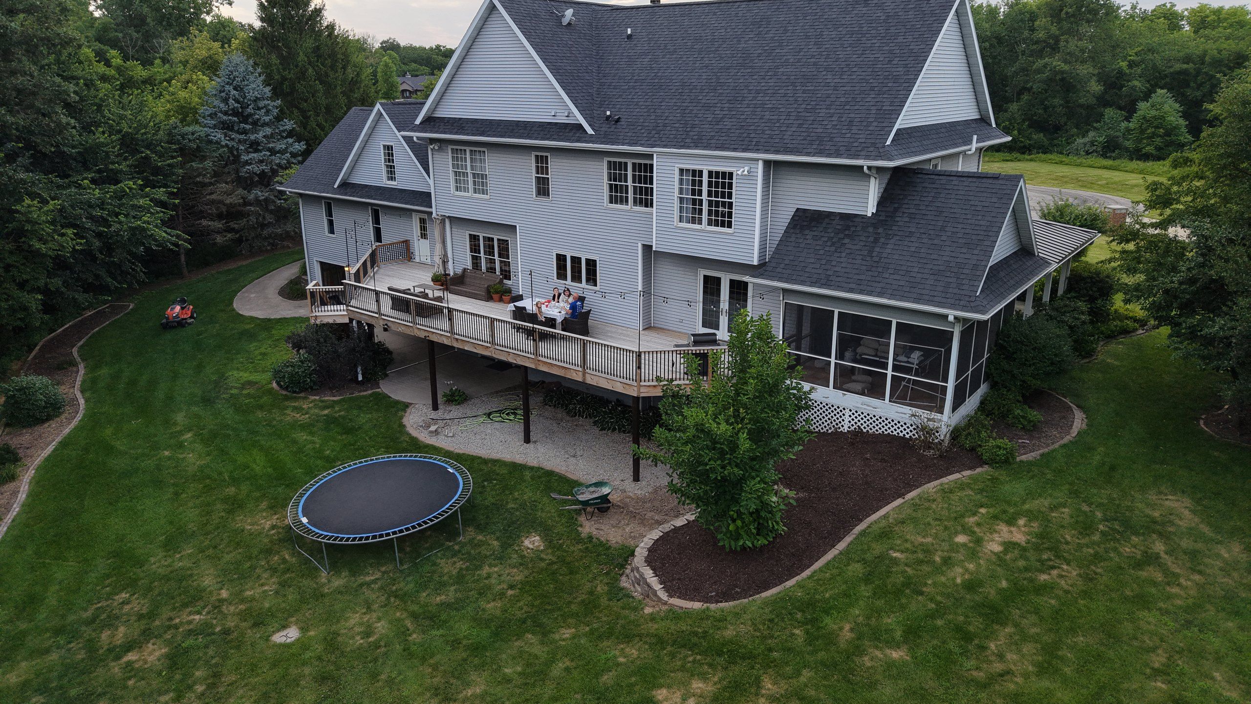 Drone photo of the property as it stands: open lawn, an old round trampoline, a wheelbarrow parked on the grass and a gravel patch under the deck. No patio, no pool, no court, no lighting.