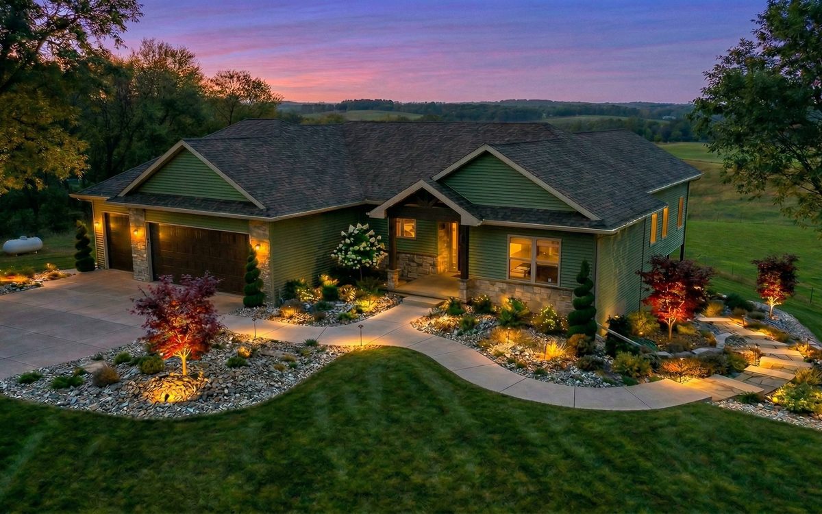 Front elevation of a ranch home at dusk with warm uplighting on maples, river-rock beds, low path lights and a lit driveway approach.