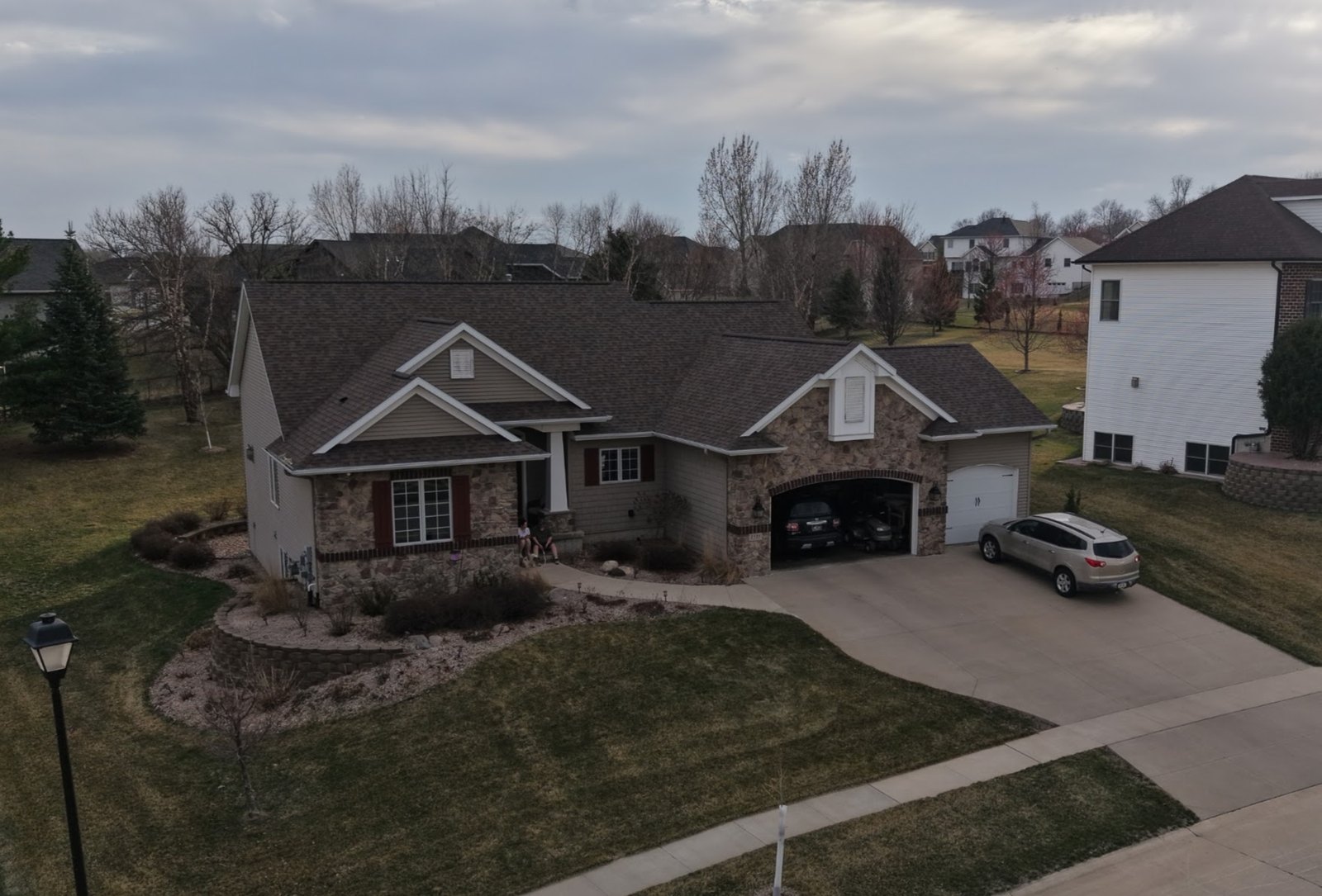 Broad curving concrete driveway sweeping to a three-car garage on a stone and shake-sided ranch home, with a concrete apron, a public sidewalk crossing the drive and terraced planting beds along the house.