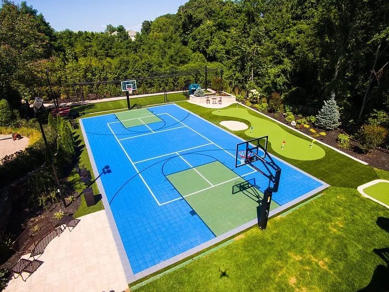 The same rear lawn from the same elevated position with a blue athletic court, green surround, white lines and a hoop at one end.