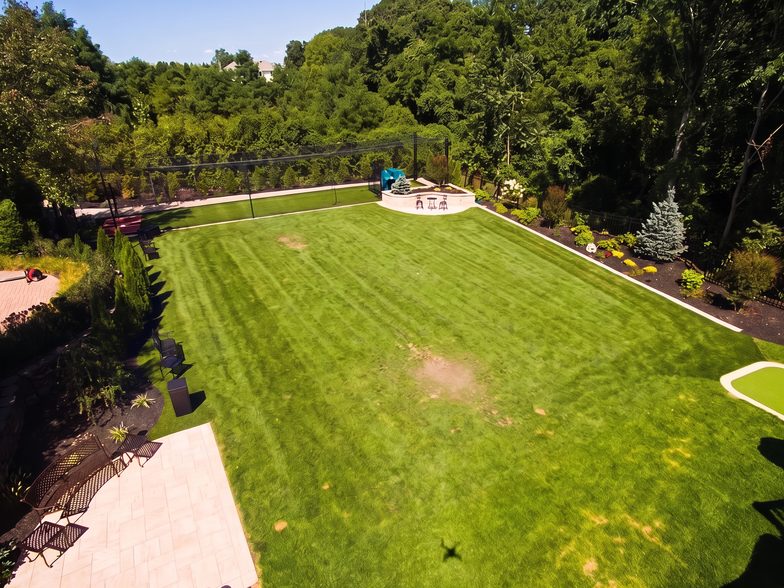 Elevated view of a wide green rear lawn edged by mature trees and a curved paver walk, with no court in place.
