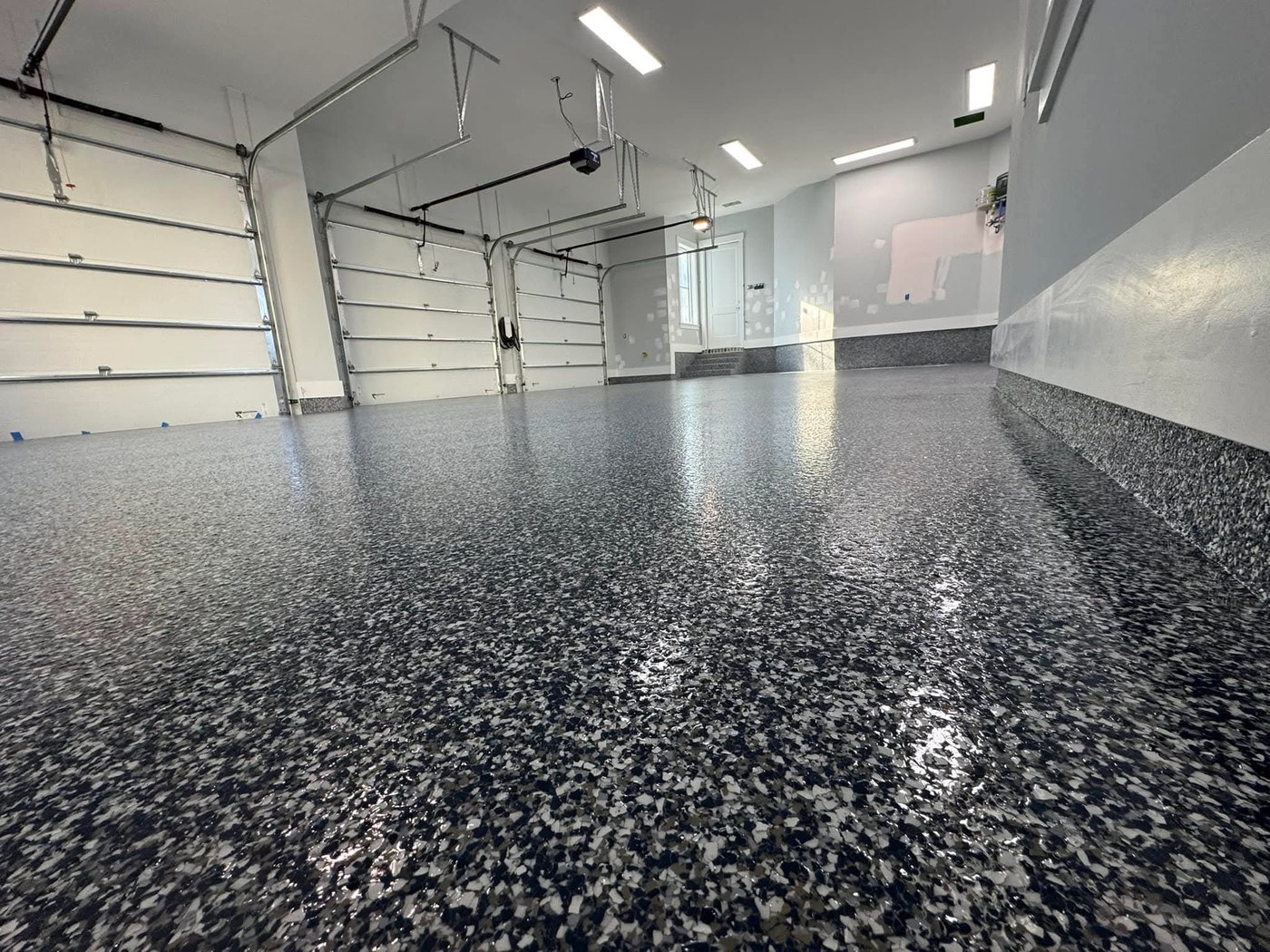 Finished grey, black and white flake epoxy floor in a multi-bay garage with white walls and a high gloss finish reflecting the overhead lights and garage doors.