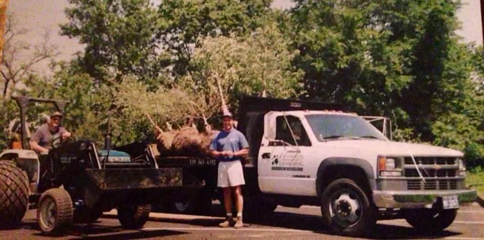 Faded 1995 photograph of two men beside a white Chamberlin Landscaping dump truck and a tractor, with a large balled and burlapped tree loaded on the flatbed.