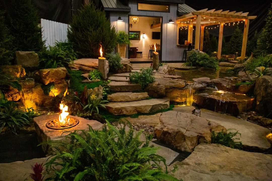 Finished landscape at dusk with warm uplighting on trees and plantings and a softly lit stone edge along the beds.