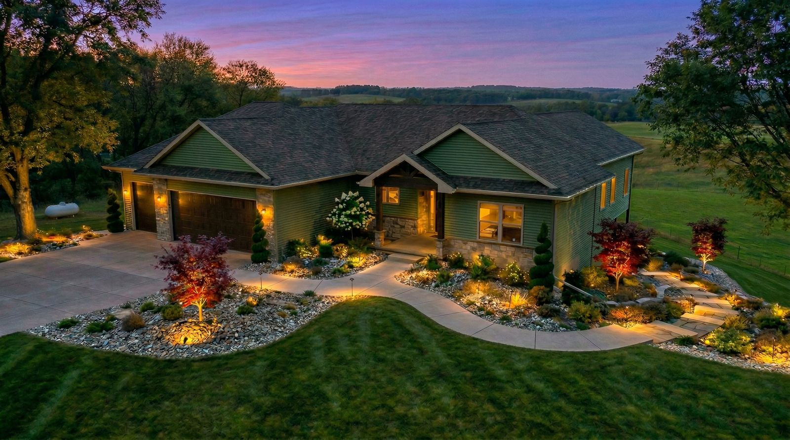 Evening photograph of a ranch home with warm uplighting in Japanese maples, lit stone entry columns, path lights along a curved walk and glowing rock beds.