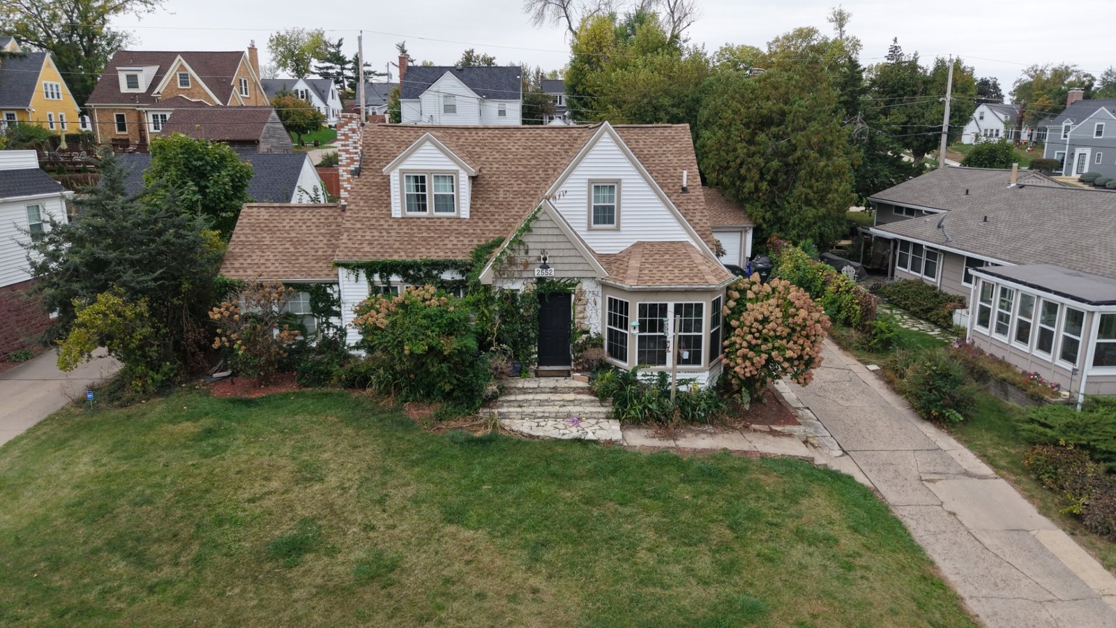 Drone view of an existing front yard and home prepared for landscape concept planning, with an open lawn, overgrown foundation plantings, a stone entry stoop and a concrete drive.