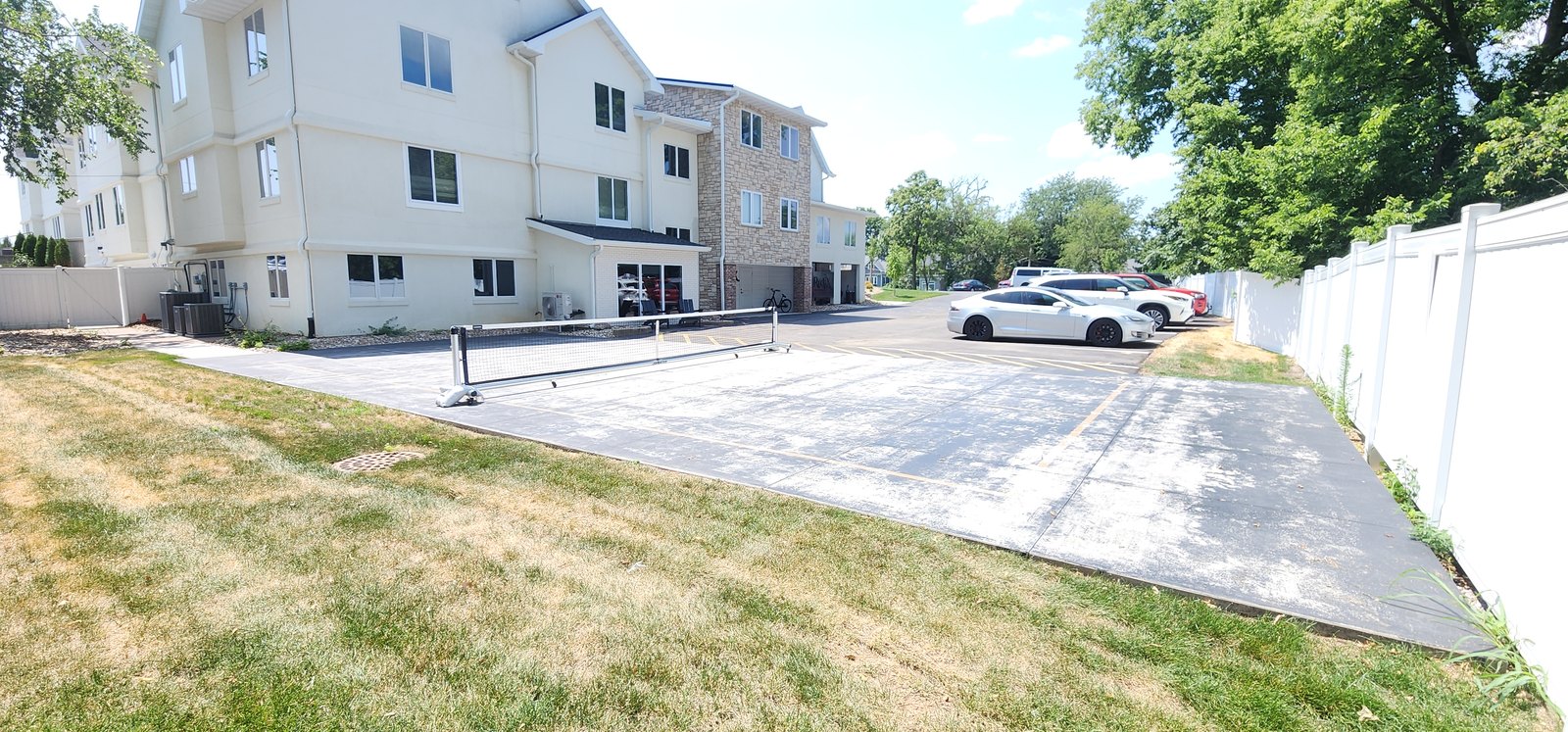 Newly placed concrete parking area and drive aisle behind a multi-unit building, bordered by a white vinyl privacy fence on one side and lawn on the other, with an asphalt lot and parked cars beyond.