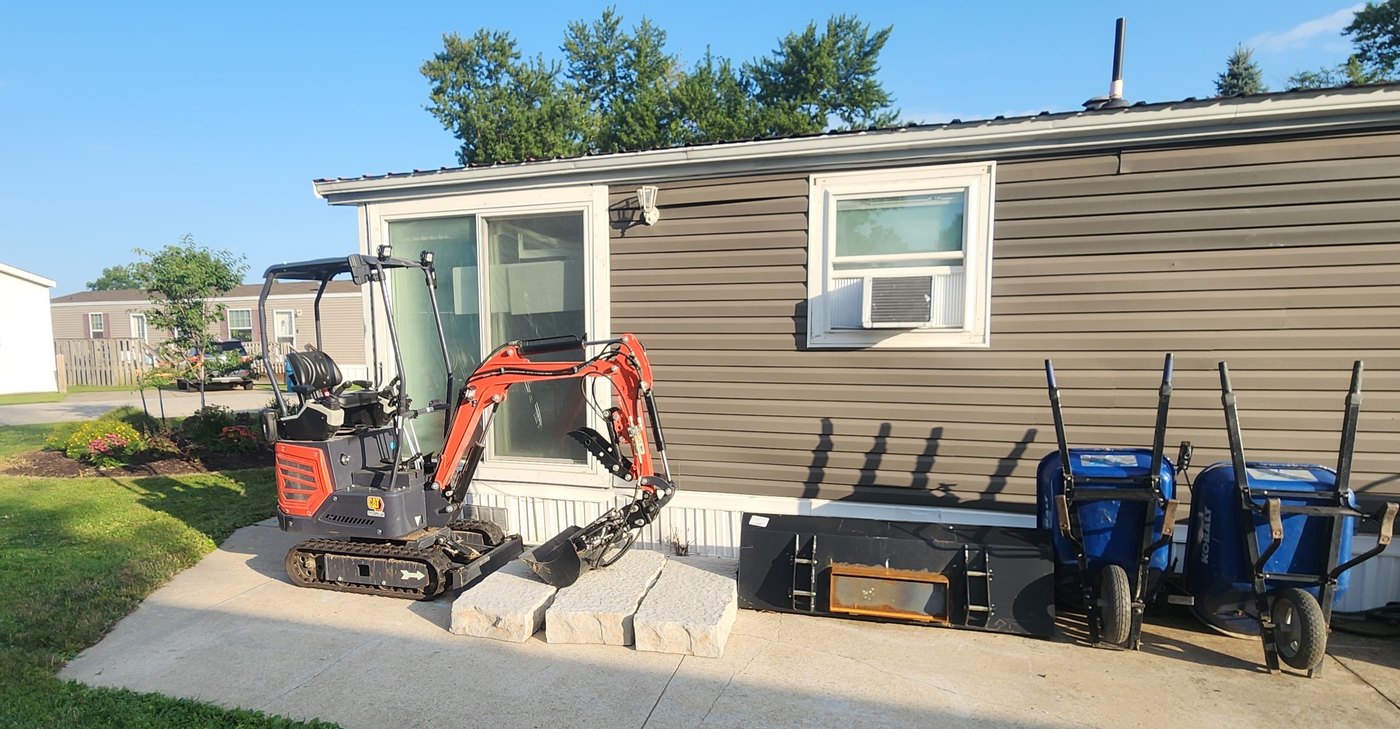 Compact tracked excavator and two concrete buggies staged on an existing concrete walk beside a home, ready for flatwork and grading.