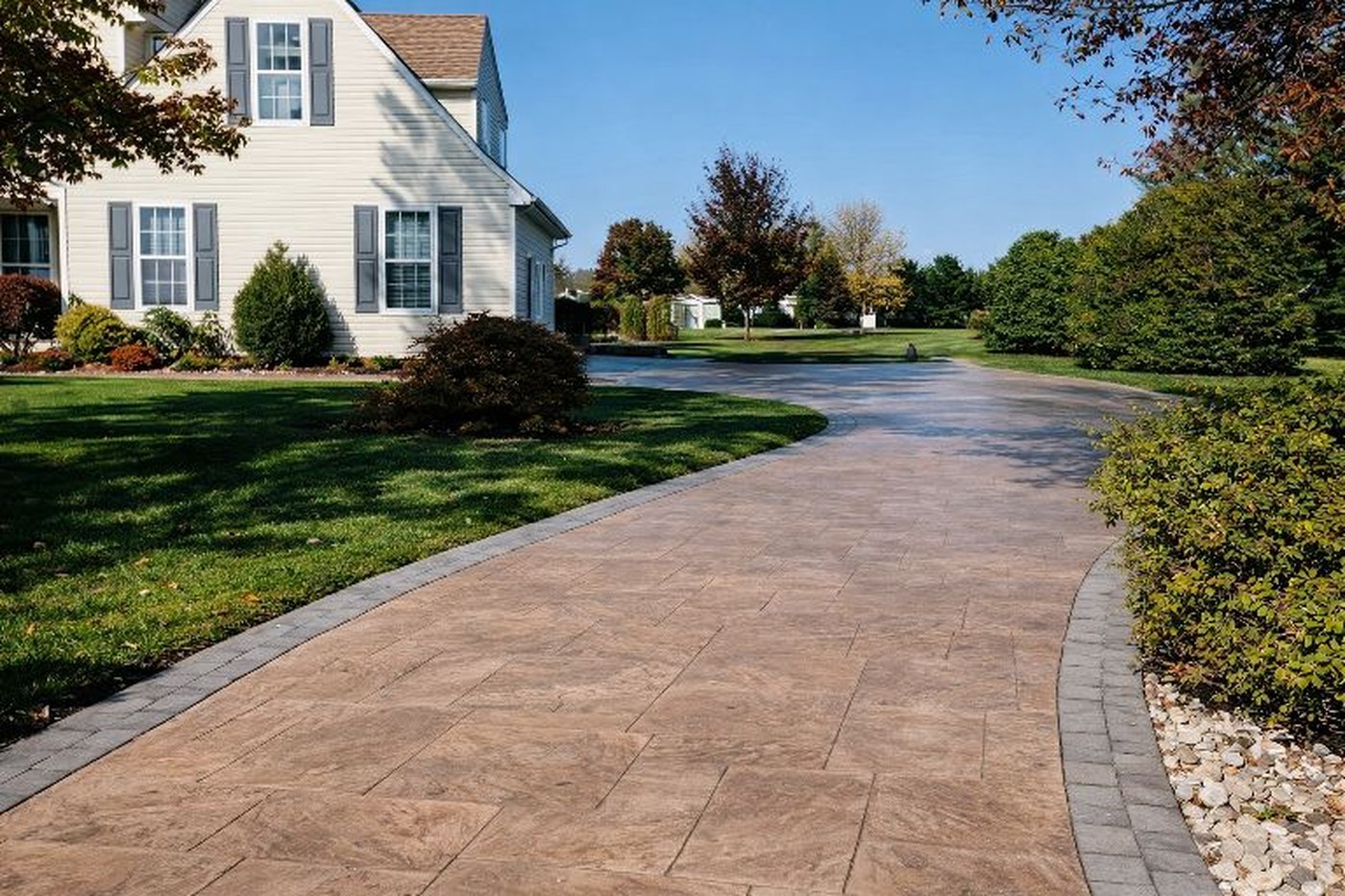 Completed stamped concrete driveway in a large tan ashlar-slate pattern, curving past a cream two-storey home, edged with a contrasting charcoal paver-look border band and river-rock bed.