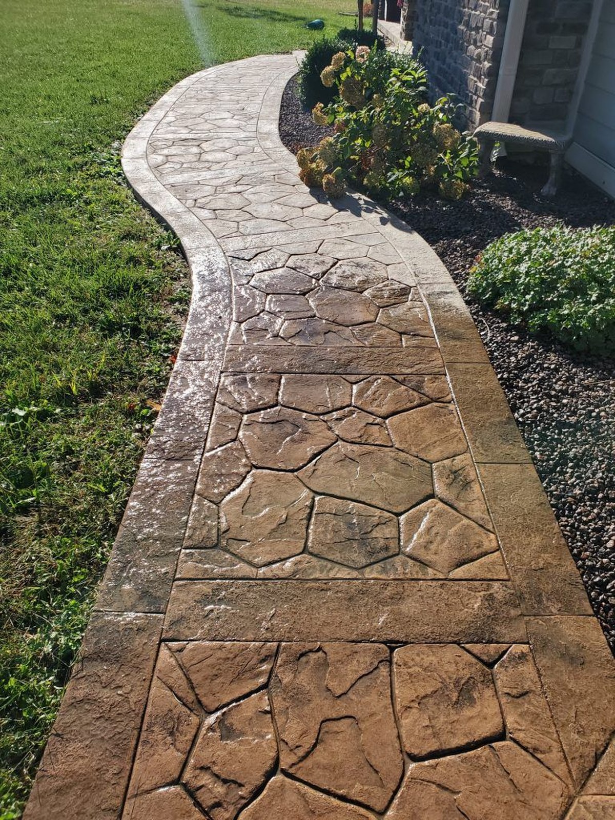 Completed curved stamped concrete walkway beside a stone-faced home: a random flagstone pattern in warm tan and brown with a smooth banded border following the curve, lawn on one side and a hydrangea bed and gravel border on the other.